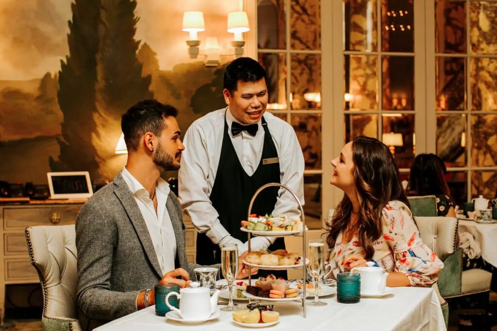 A waiter in a black vest and bow tie serving a three-tiered platter of afternoon tea treats to a smiling couple seated at a table, with holiday decorations visible in the background.