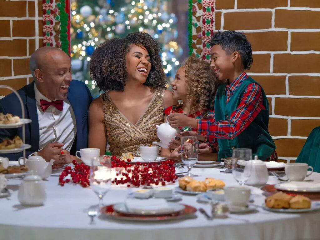 A smiling family of four, including two parents and two children, sitting around a festive white table enjoying holiday dining. The background features a large gingerbread house decoration.