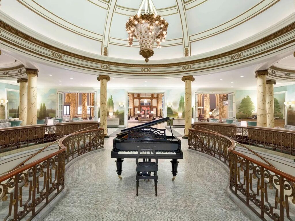 A grand interior view of the Laurel Court's rotunda, featuring a black grand piano centered on a mosaic floor, surrounded by a curving balustrade, ionic columns, and a high dome with a crystal chandelier.