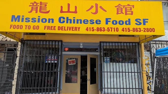 Exterior street view of the Mission Chinese Food SF restaurant entrance, featuring a bright yellow awning with the restaurant's name in English and Chinese characters. The restaurant is closed with a metal security gate. A lively spot for a meal during Christmas In San Francisco.