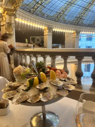 A seafood platter with oysters, shrimp, and lemon on a table inside The Rotunda at Neiman Marcus, showing the beautiful glass dome and golden accents, suggesting a luxury dining experience for Christmas In San Francisco.