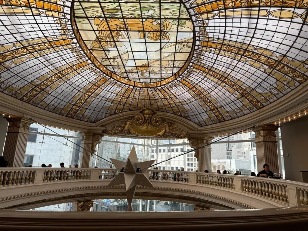 A grand interior view of The Rotunda restaurant at Neiman Marcus, highlighting the ornate gold trim and the striking stained-glass ceiling, with a table set in the foreground,