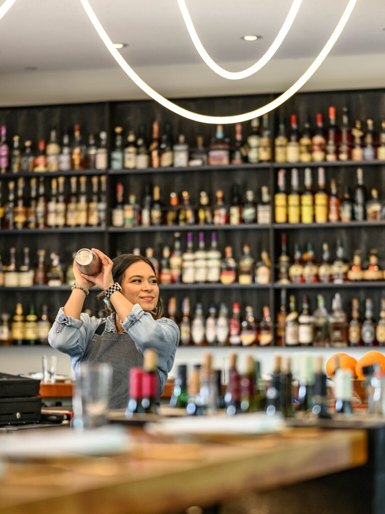 Bartender shaking a cocktail shaker behind a well-stocked bar with glowing overhead lighting.