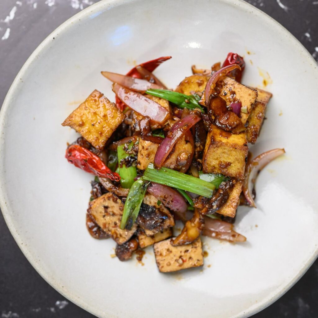 A close-up of a dish featuring stir-fried tofu pieces, red onion slices, green onions, and dried red chili peppers in a dark sauce, served in a white bowl.