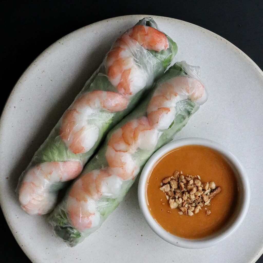 Two fresh shrimp and vegetable spring rolls with a side of peanut dipping sauce topped with crushed peanuts on a white plate.