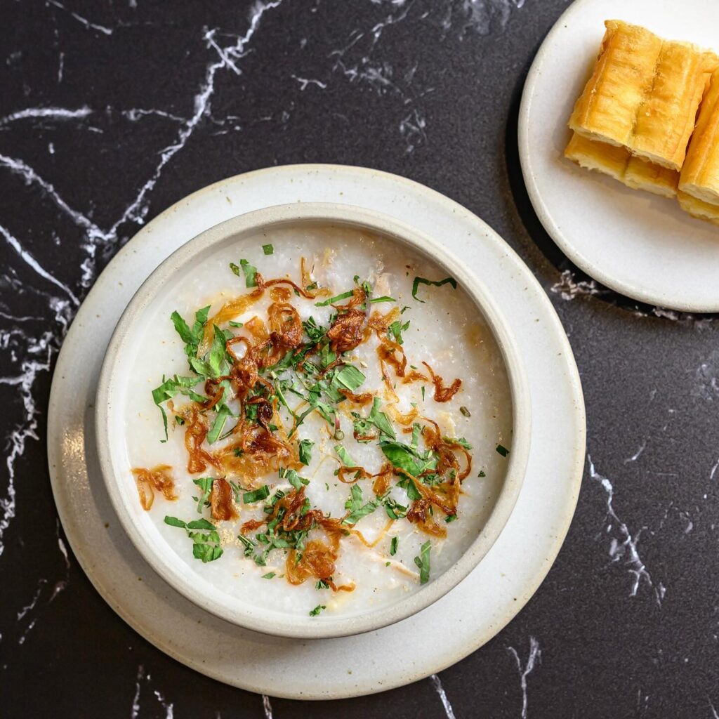 A bowl of Vietnamese chicken congee (cháo gà) topped with crispy fried shallots and herbs, served next to two pieces of savory fried dough (youtiao) on a black marble surface.