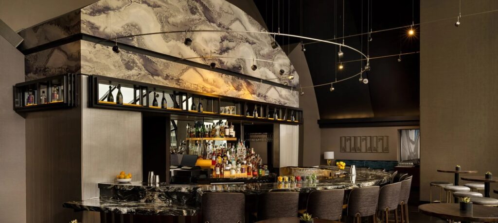 A modern and sophisticated bar area with a curved, dark marble countertop, high-backed stools, an arched wall with a marbled finish, and bottles displayed on shelving.