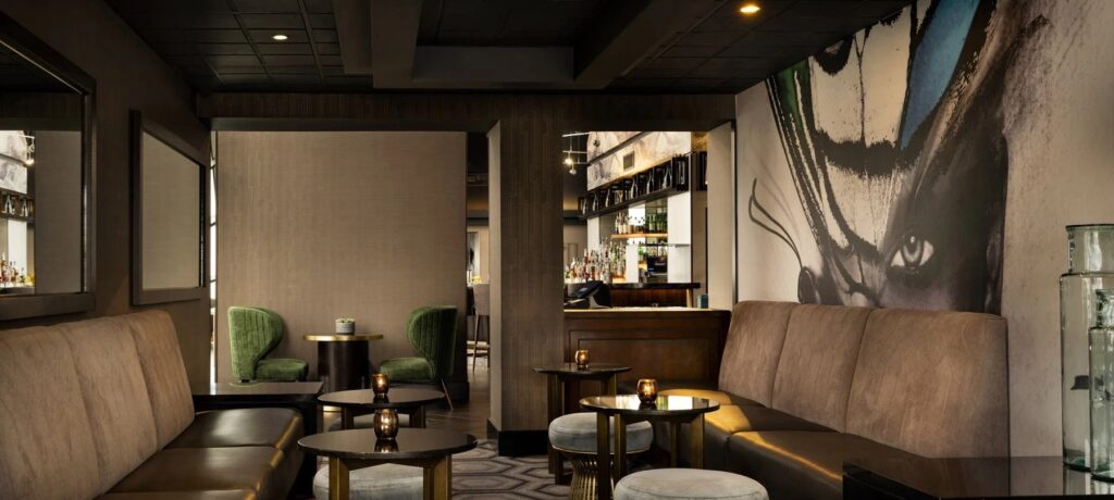 A cozy seating area in a lounge with light brown upholstered couches, two small green accent chairs, and a partial view of the bar area in the background.