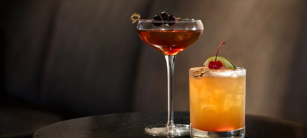 Two cocktails on a table: one dark cocktail in a coupe glass garnished with two dark cherries, and one light-colored, tall cocktail in a rocks glass garnished with a lime wedge and a bright red cherry.