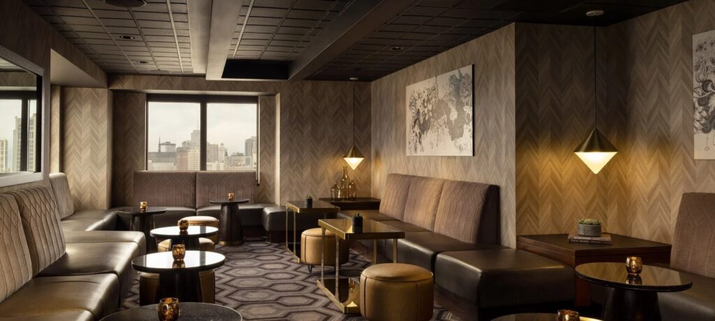 An interior shot of a stylish, modern lounge area featuring low, plush leather seating in a taupe color, dark round tables, and a geometric patterned carpet.