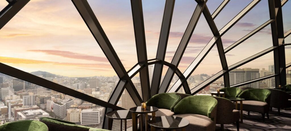A stunning panoramic view of a city skyline and distant hills at sunset, seen through the large, geometrically-framed windows of a modern lounge with green velvet armchairs.