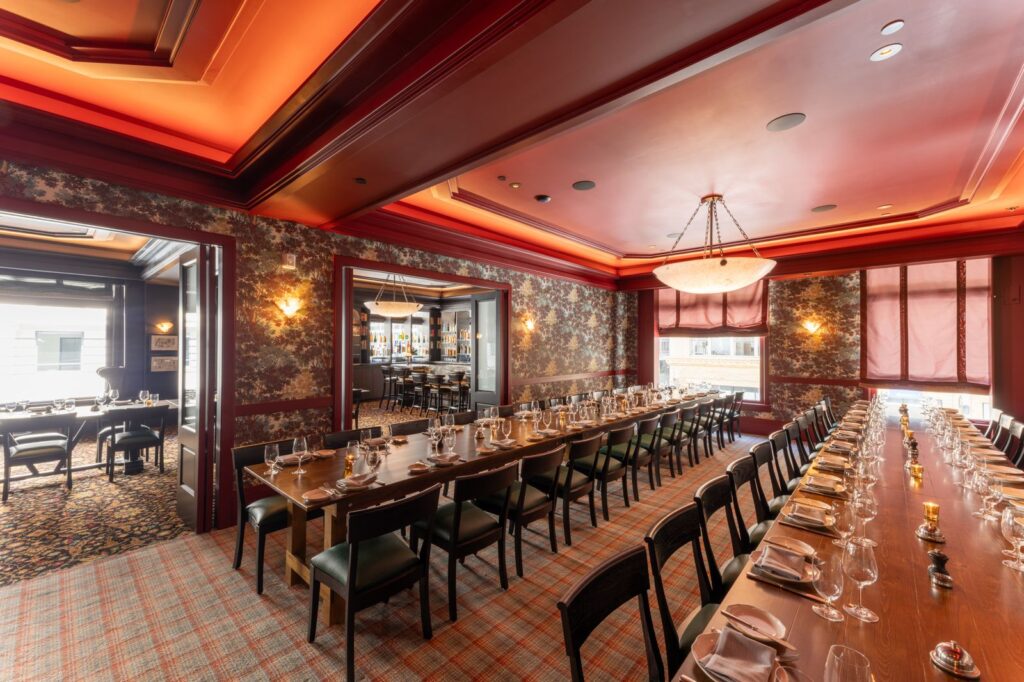 Long, formal dining room in deep red and wood tones, set with long tables for a large private event. The ceiling and walls have rich textures and lighting, suggesting a luxurious holiday dinner setting for Christmas in San Francisco.