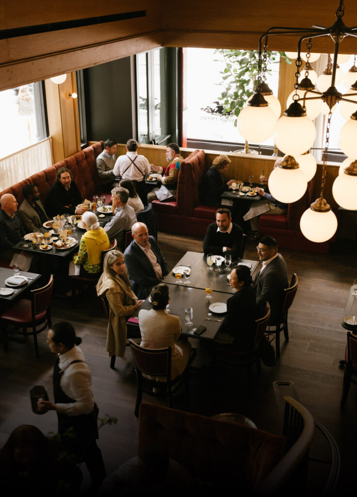 An aerial view of a lively restaurant dining area with patrons seated in plush, red velvet booths and at dark tables. The space has warm lighting and large windows, capturing a busy holiday lunch atmosphere during Christmas in San Francisco.