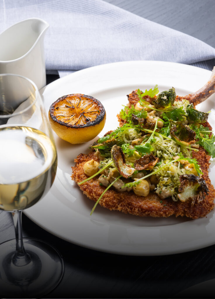 Close-up of a golden-brown, breaded pork chop (or schnitzel) topped with a salad of greens and roasted vegetables, served on a white plate with a charred lemon half and a glass of white wine. A festive, upscale dining experience for Christmas in San Francisco.