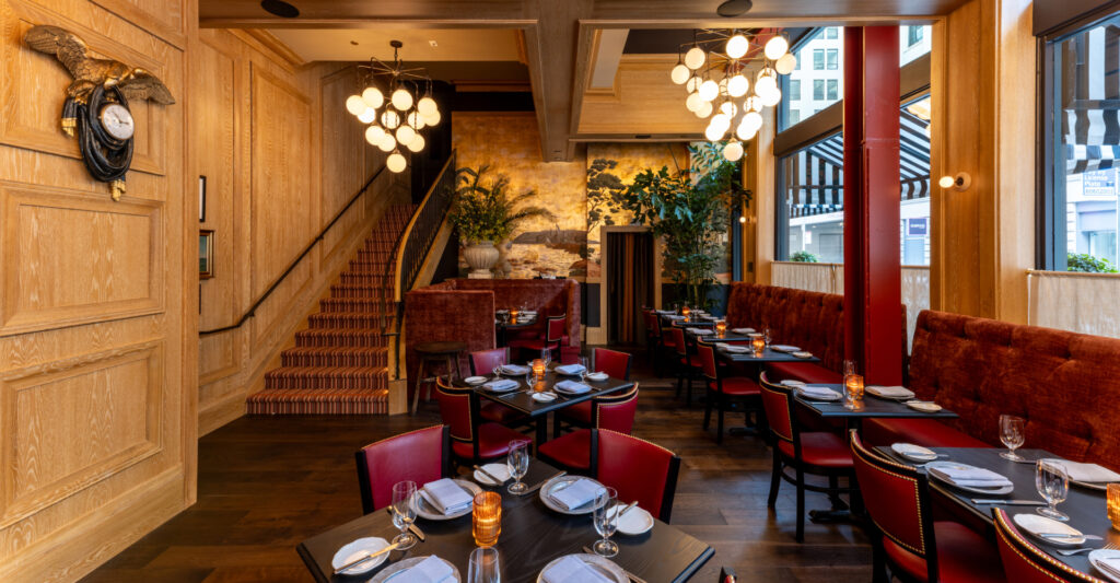 A warm, inviting restaurant interior with red velvet seating, dark tables, light wood paneling, and a grand staircase covered in a striped runner. The sophisticated decor creates an upscale and cozy ambiance for Christmas in San Francisco dining.
