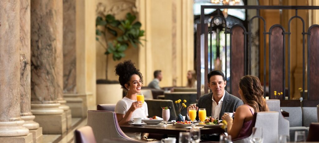 Three well-dressed people enjoying breakfast at a table in the Garden Court, with large, classic marble columns visible in the background. One woman is smiling while holding a glass of orange juice.