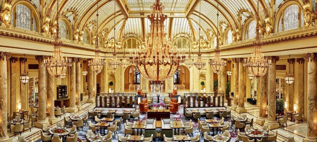A breathtaking, wide-angle shot of the entire Garden Court, highlighting the immense arched glass ceiling and the enormous central crystal chandelier. The space is set up with many tables for dining.