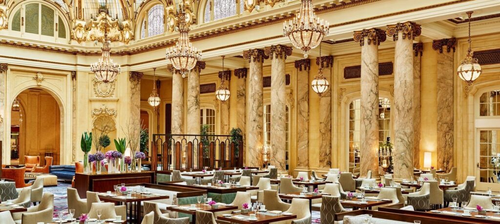 The grand, opulent interior of the Garden Court restaurant, featuring towering marble columns, richly gilded ornamentation, and multiple sparkling crystal chandeliers over set dining tables.