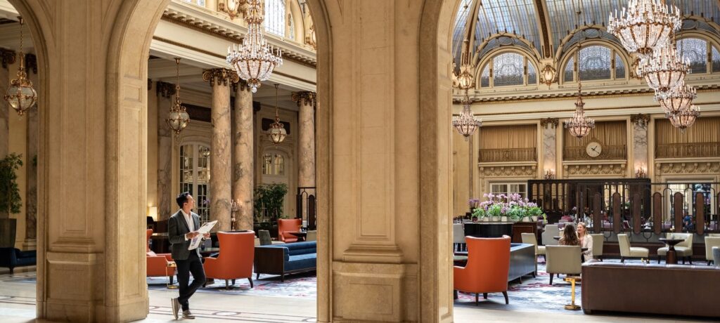A full-length view of the Garden Court's stunning architecture, showing a man walking through a stone archway while holding a newspaper. The space is filled with high arches, crystal chandeliers, and a mix of dining and lounge seating.