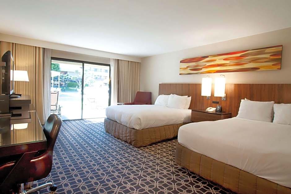 A bright guest room at Hilton Palm Springs with two queen beds, modern wood accents, and a sliding glass door leading to a balcony overlooking the Splash House 2026 festivities.