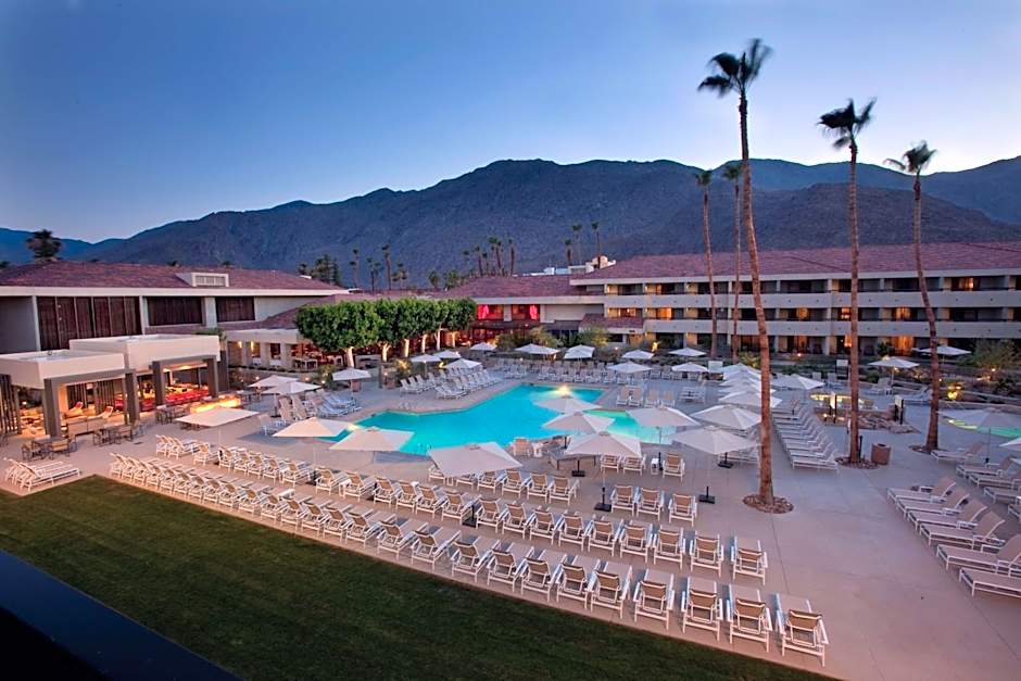 An expansive aerial view of the Hilton Palm Springs pool deck and resort buildings against the San Jacinto mountains, serving as a primary host venue for the Splash House 2026 pool parties.