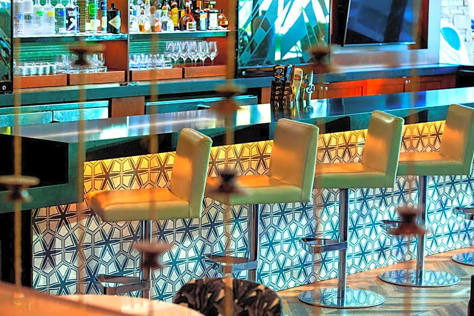 A close-up of a stylish hotel bar with lime-green leather barstools. The bar front is decorated with a blue and white geometric tile pattern, and the back bar is stocked with various spirits and glassware under warm yellow accent lighting.