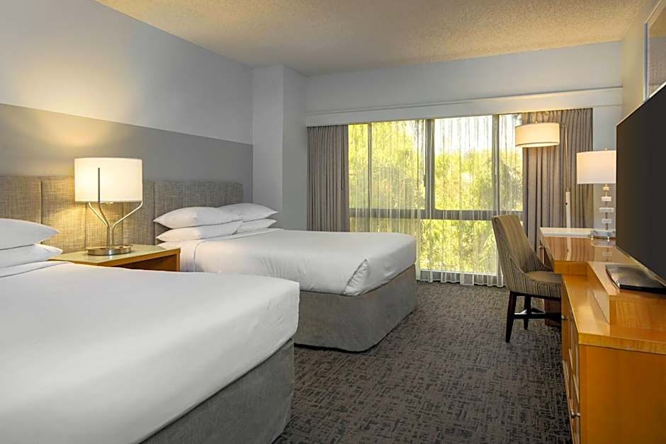 A clean, contemporary hotel room with two queen beds featuring white linens and gray padded headboards. The room includes a wooden work desk with a lamp and a flat-screen TV on a dresser, with sheer curtains covering a large window.