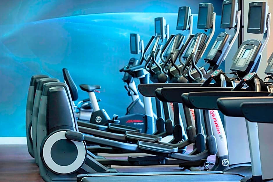 An indoor hotel gym featuring a row of modern Life Fitness treadmills and elliptical machines. The equipment faces a vibrant blue wall with an abstract wave design, and large windows are visible in the background.