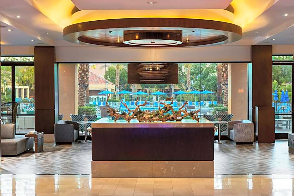 A spacious, modern hotel lobby featuring a large dark wood central counter topped with a decorative driftwood sculpture. The room has high ceilings with a circular light fixture, herringbone-patterned floors, and floor-to-ceiling windows looking out toward a pool area with blue umbrellas.