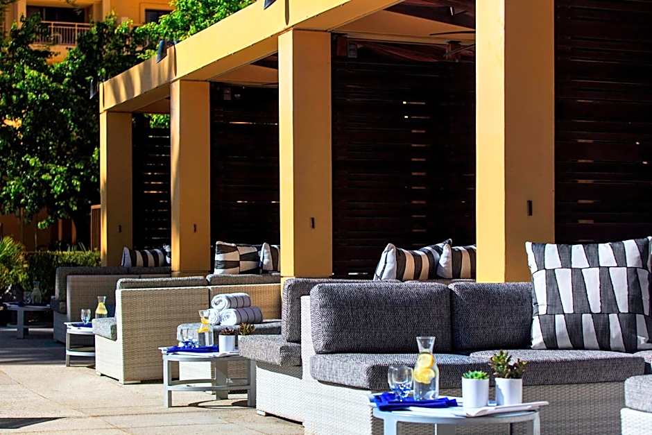 A sunny outdoor lounge area featuring gray wicker sofas with black and white patterned pillows. Each seating section is shaded by a yellow architectural pergola and includes small side tables with water carafes and potted succulents.