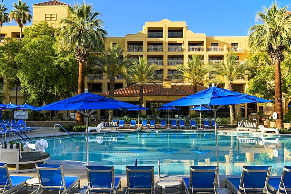 A large outdoor swimming pool surrounded by bright blue lounge chairs and matching umbrellas. The Renaissance Palm Springs Hotel building, featuring desert-style architecture and palm trees, serves as the backdrop under a clear blue sky.