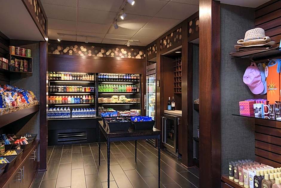 A well-lit hotel marketplace or gift shop. Dark wood shelves and refrigerated coolers are stocked with snacks, bottled drinks, and wine. A side wall displays retail items like sun hats, beach bags, and sunscreens.