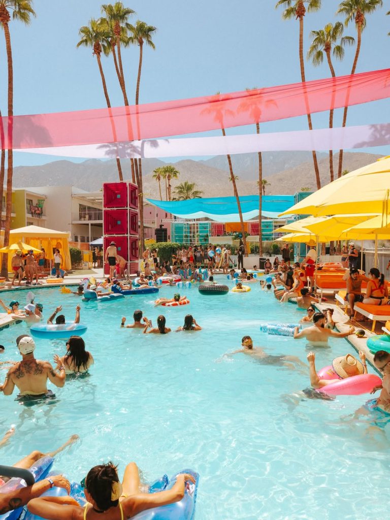 A vibrant, wide-angle shot of a crowded pool party at Splash House 2026 in Palm Springs, featuring attendees on floats, yellow umbrellas, and pink decorative streamers against a desert mountain backdrop.