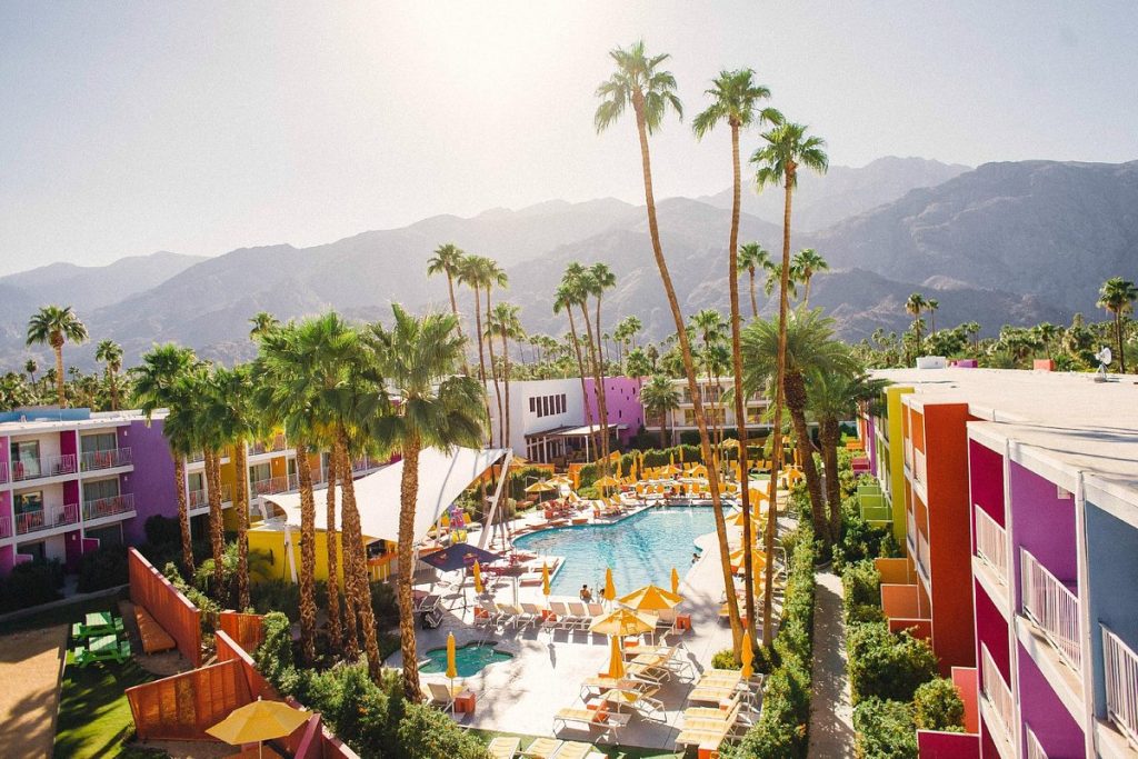 A high-angle, wide shot of a vibrant desert resort featuring a sparkling blue pool surrounded by bright yellow umbrellas and lounge chairs. The hotel buildings are painted in a rainbow of saturated colors, set against a backdrop of tall palm trees and rugged, hazy mountains.