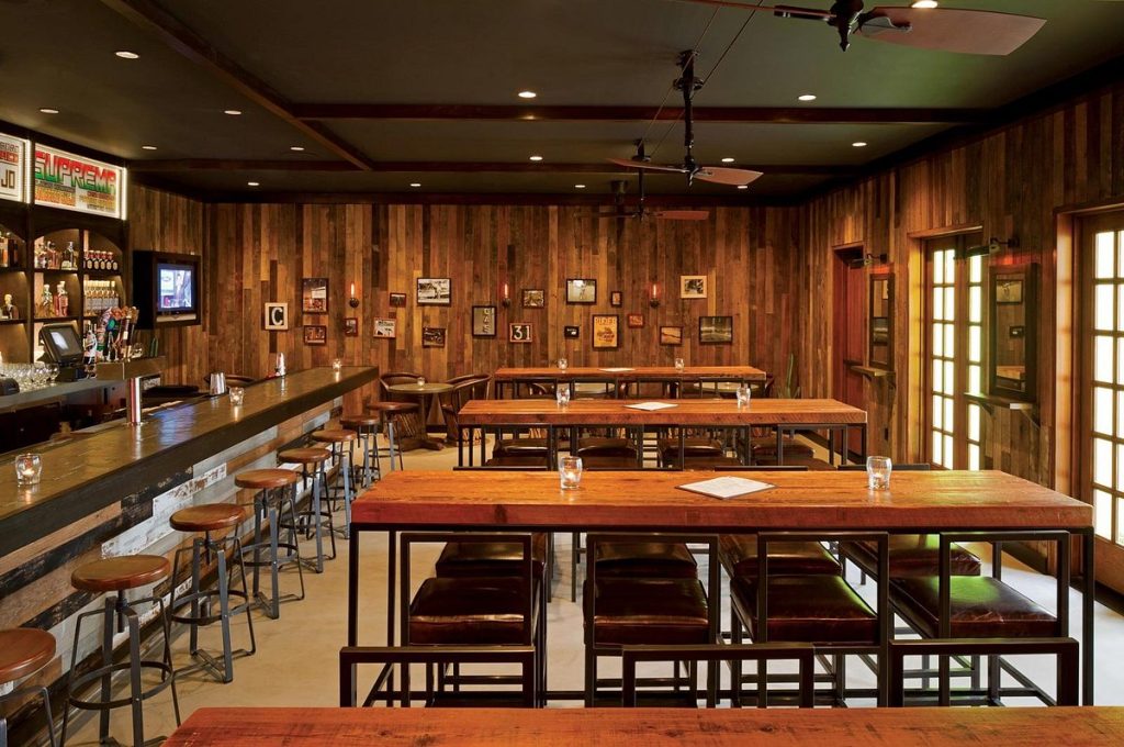 A spacious, modern lobby featuring a bright purple ping-pong table in the center. The room includes a curved reception desk with a loud floral backdrop, leather lounge seating, and a "Tequila" neon sign visible in the adjacent bar area.