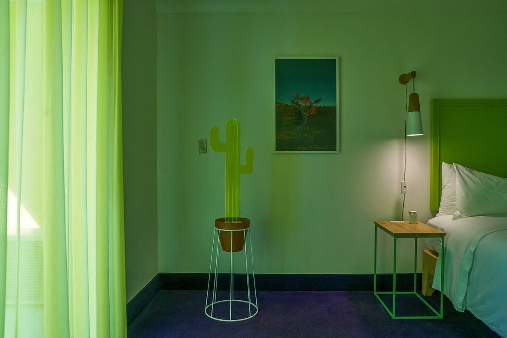 A minimalist detail shot of a guest room showing a lime-green headboard and matching sheer curtains. A stylized yellow cactus silhouette in a pot sits on a wire stand next to a simple wood and metal nightstand.