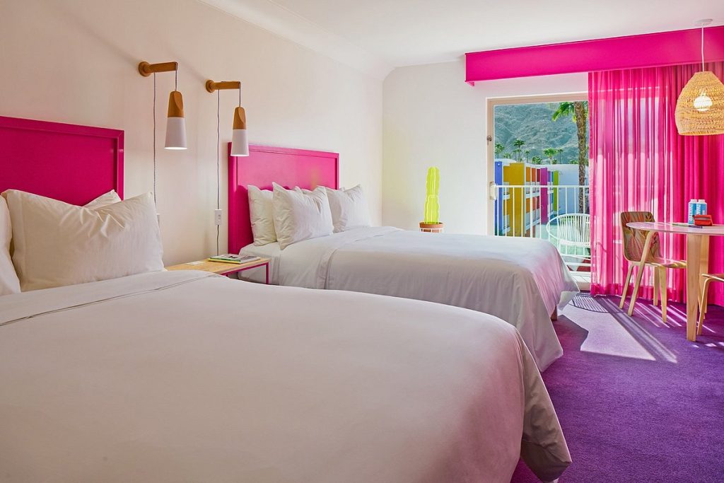 A bright hotel room with two queen beds, featuring a bold hot-pink headboard and matching pink blackout curtains. A neon lime-green cactus lamp stands in the corner, and a private balcony offers a view of the colorful hotel exterior and mountains.