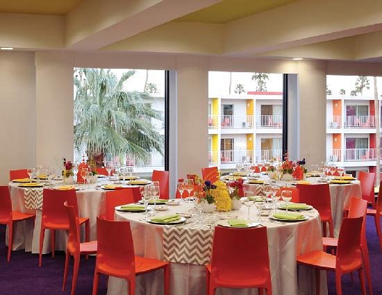 An indoor event space set for a banquet with round tables, white linens, and bright orange chairs. Large windows look out onto the colorful hotel courtyard, while the purple carpeting and chevron-patterned table runners add to the playful decor.