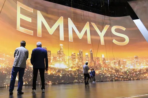 Crew members and production staff stand on stage in front of a large illuminated backdrop displaying the word "EMMYS" above a glowing cityscape, likely during preparations or rehearsals for the Emmy Awards