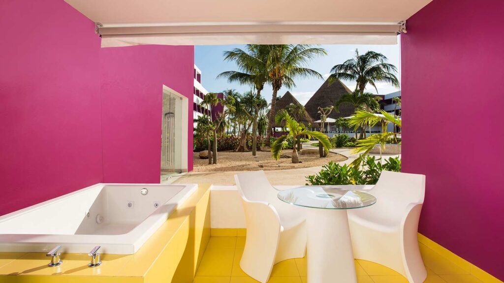 A brightly colored hotel room patio with magenta walls and a yellow floor. A white, rectangular hot tub is on the left, and a small white table with two chairs is on the right, overlooking a tropical outdoor area with palm trees.
