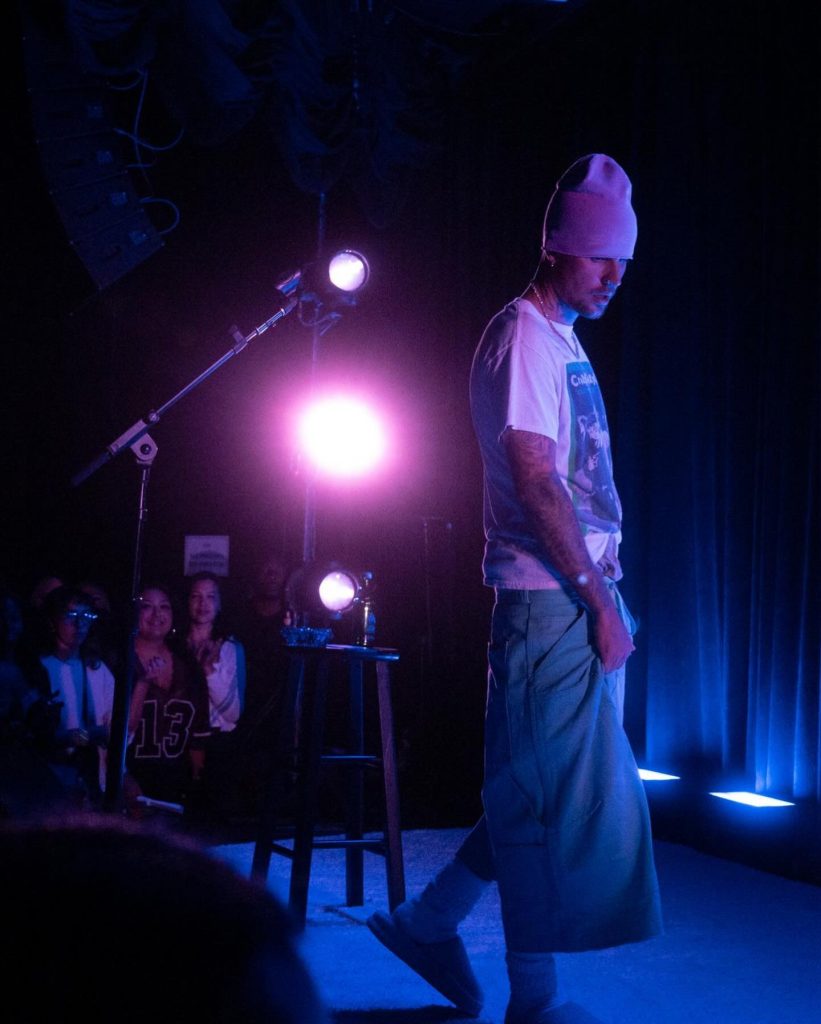 Justin Bieber performing on a dimly lit stage under vibrant purple and blue spotlights. He is wearing a pink beanie, a white graphic t-shirt, and oversized teal trousers. In the background, a small audience is visible, and the scene captures the atmosphere surrounding the Coachella 2026 Bieber setlist leak that has stunned fans.