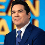 A medium close-up photo of actor Dean Cain. He is smiling slightly and looking off to the side while wearing a navy blue suit, a white collared shirt, and a patterned blue tie.