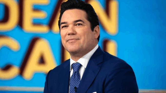 A medium close-up photo of actor Dean Cain. He is smiling slightly and looking off to the side while wearing a navy blue suit, a white collared shirt, and a patterned blue tie.