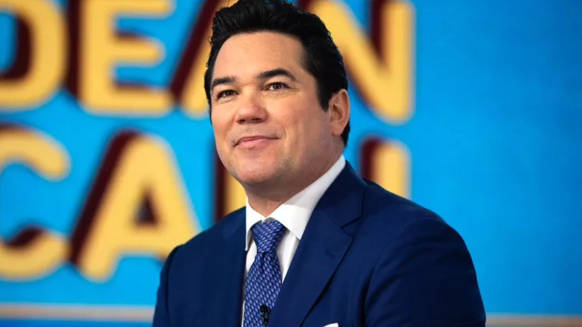A medium close-up photo of actor Dean Cain. He is smiling slightly and looking off to the side while wearing a navy blue suit, a white collared shirt, and a patterned blue tie.
