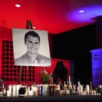 A night photo of a vigil with a large portrait of Charlie Kirk on an easel, surrounded by dozens of lit candles, flowers, and small American flags, under dramatic red and blue lighting. Charlie Kirk Beliefs.