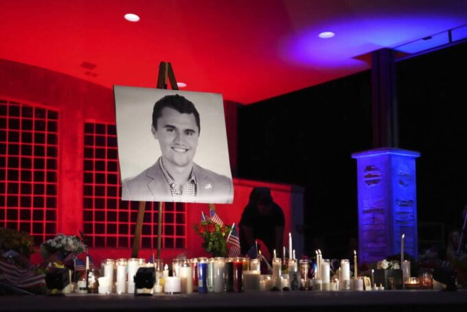 A night photo of a vigil with a large portrait of Charlie Kirk on an easel, surrounded by dozens of lit candles, flowers, and small American flags, under dramatic red and blue lighting. Charlie Kirk Beliefs.