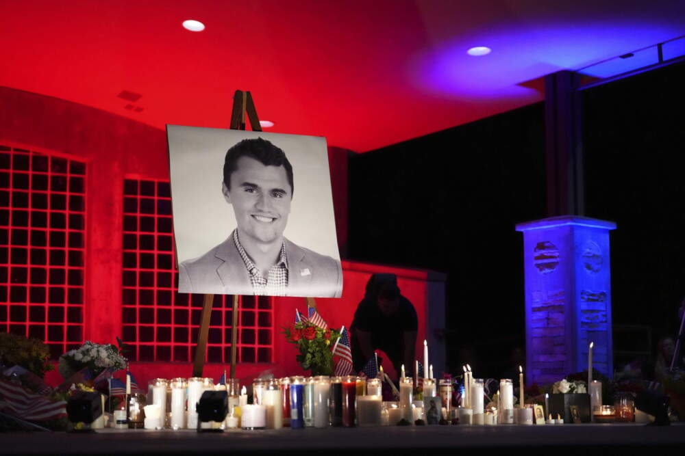 A night photo of a vigil with a large portrait of Charlie Kirk on an easel, surrounded by dozens of lit candles, flowers, and small American flags, under dramatic red and blue lighting. Charlie Kirk Beliefs.