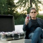 Actor Charlie Sheen sitting outdoors next to a briefcase filled with cans of his Non Alcoholic Beer brand, Wild AF Brewing. He is holding one can up and looking at the camera.