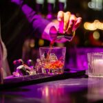 A bartender in a purple-lit environment is pouring a reddish liquid from a jigger into a glass filled with ice and a red garnish. To the right, two other glasses filled with white and pink liquids are visible, with out-of-focus lights in the background.