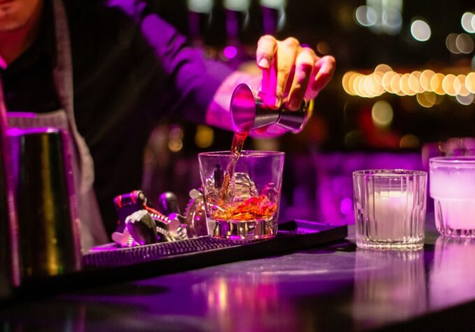 A bartender in a purple-lit environment is pouring a reddish liquid from a jigger into a glass filled with ice and a red garnish. To the right, two other glasses filled with white and pink liquids are visible, with out-of-focus lights in the background.